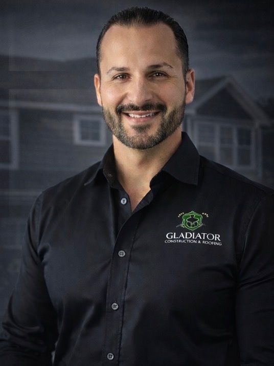 Smiling man in a black button-down shirt with a Gladiator logo, standing against a blurred house backdrop