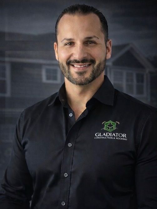 Smiling man in a black button-down shirt with a Gladiator logo, standing against a blurred house backdrop