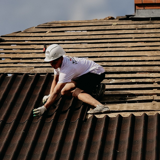 Worker in hard hat climbing a steep roof, securing dark shingles under bright sunlight