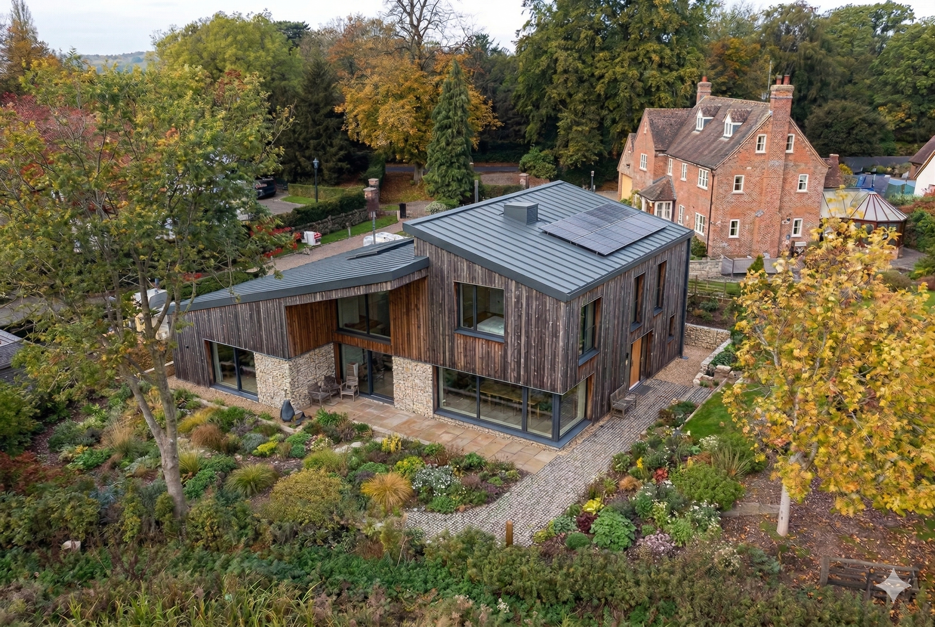 Modern two-story house with dark wood cladding, stone accents, and a slanted roof, set in a lush, landscaped garden.