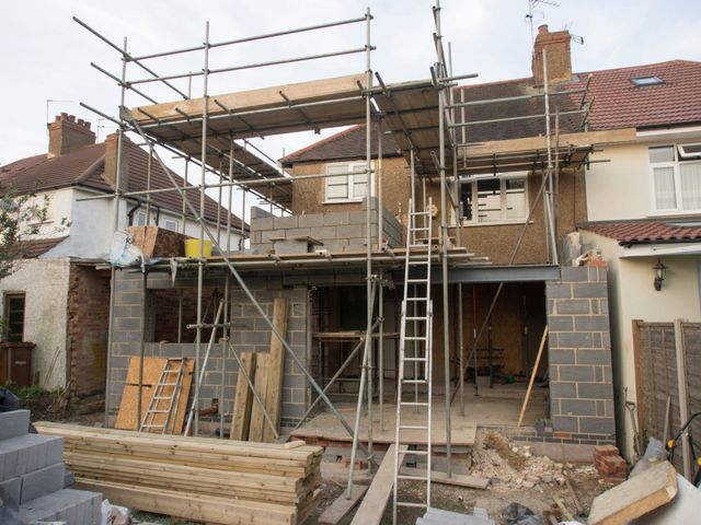 A residential construction site showing a two-story house extension in progress with scaffolding and concrete block walls.