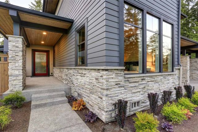A modern home entrance with a stone-clad base, dark gray lap siding, large windows, and a concrete path to the front door.