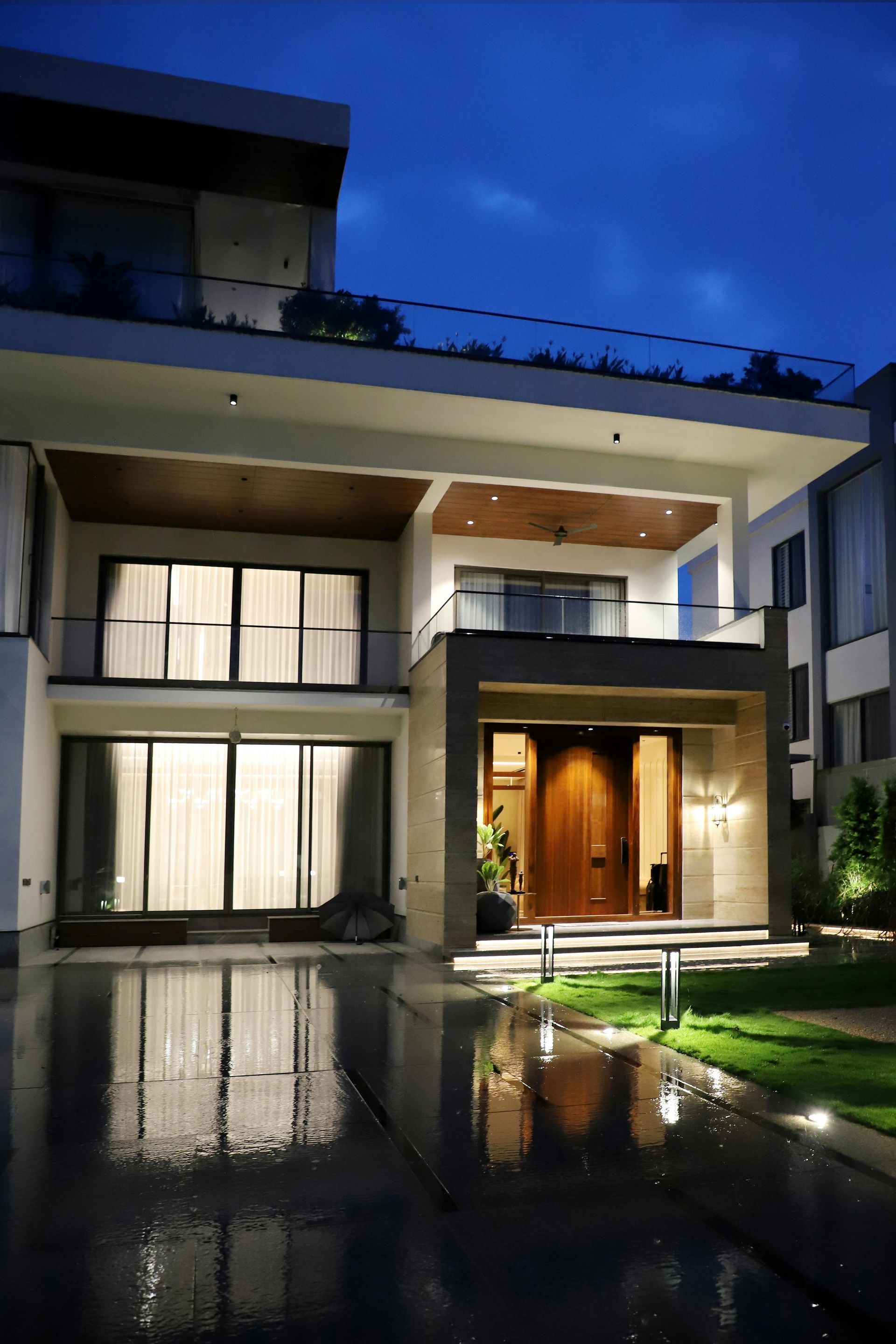A modern two-story house at night, featuring warm interior lighting, a grand wooden door, and a reflecting pool in front.