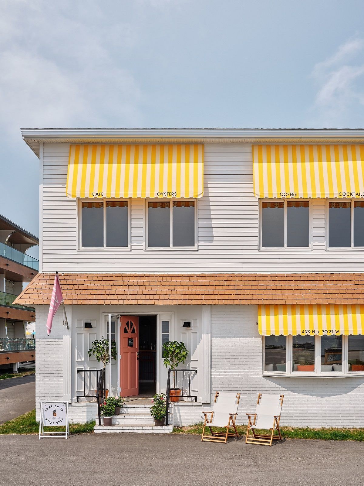 A white two-story building with yellow striped awnings, a red front door, and two lounge chairs in front.