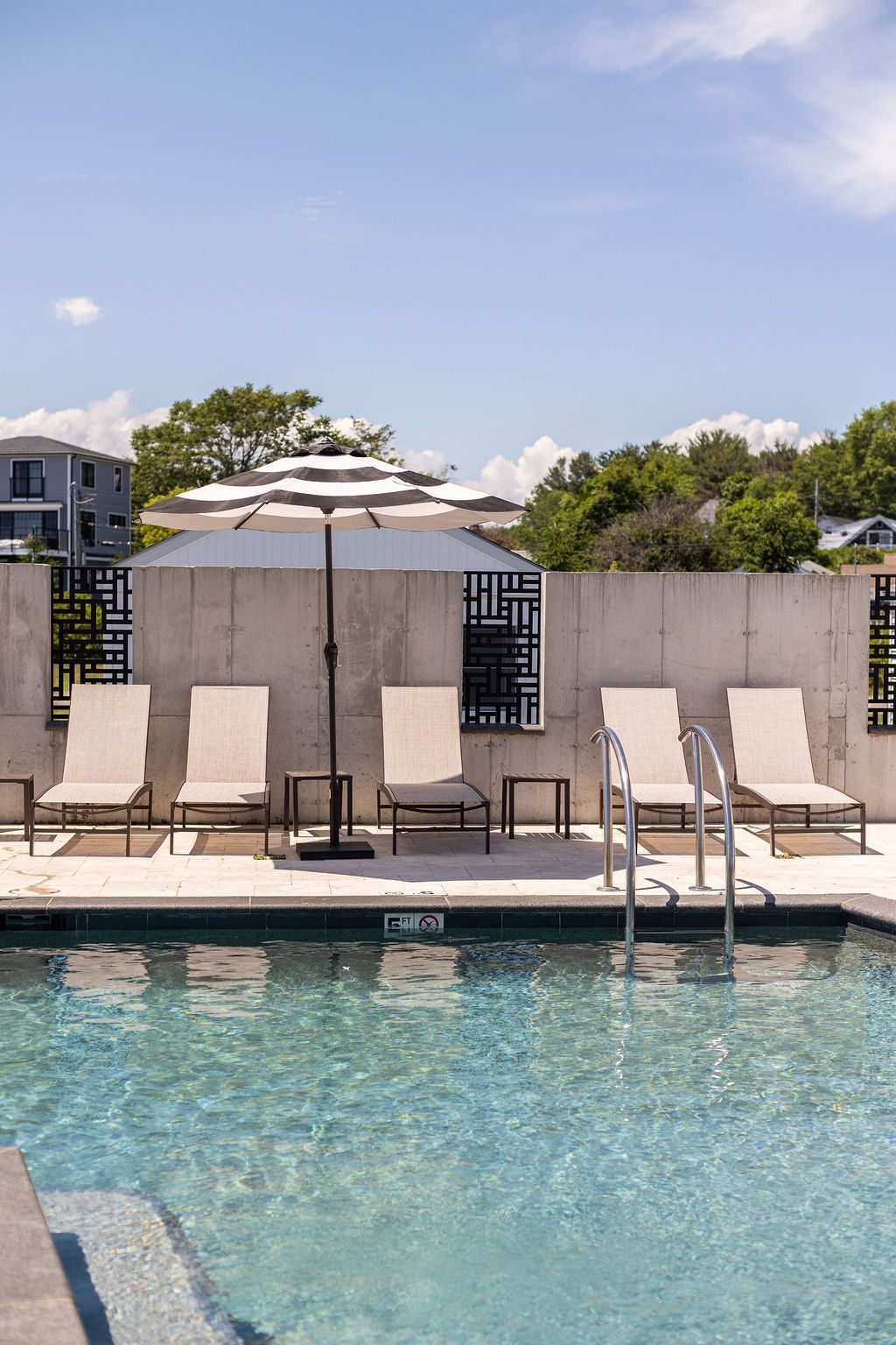 Poolside lounge chairs with colorful striped cushions sit under yellow and teal-white umbrellas against a white wall.