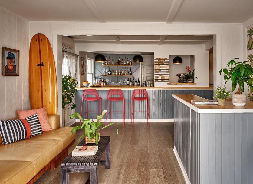 A modern, beach-themed living room with a tan sofa, surfboard, coffee table, and a bar area with three red stools.