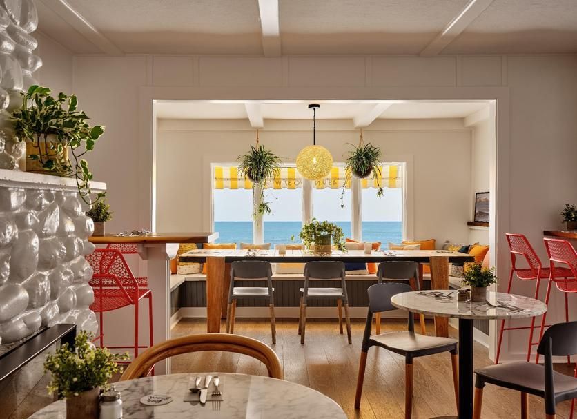 A bright, airy restaurant dining area with tables, chairs, and a long bench looking out at the ocean through large windows.