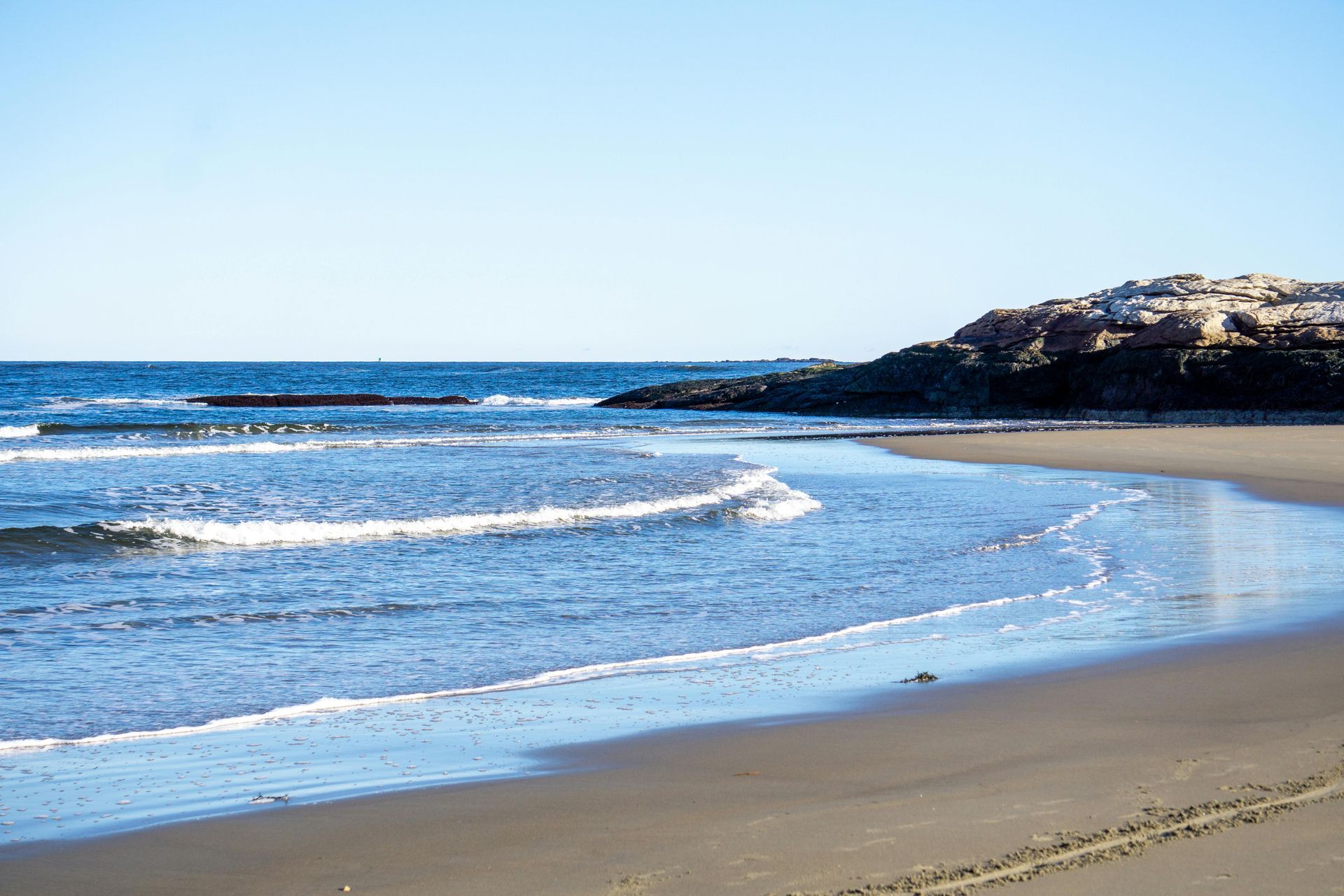 A calm, sandy beach with light waves gently lapping the shore, bordered by dark coastal rocks under a clear blue sky.