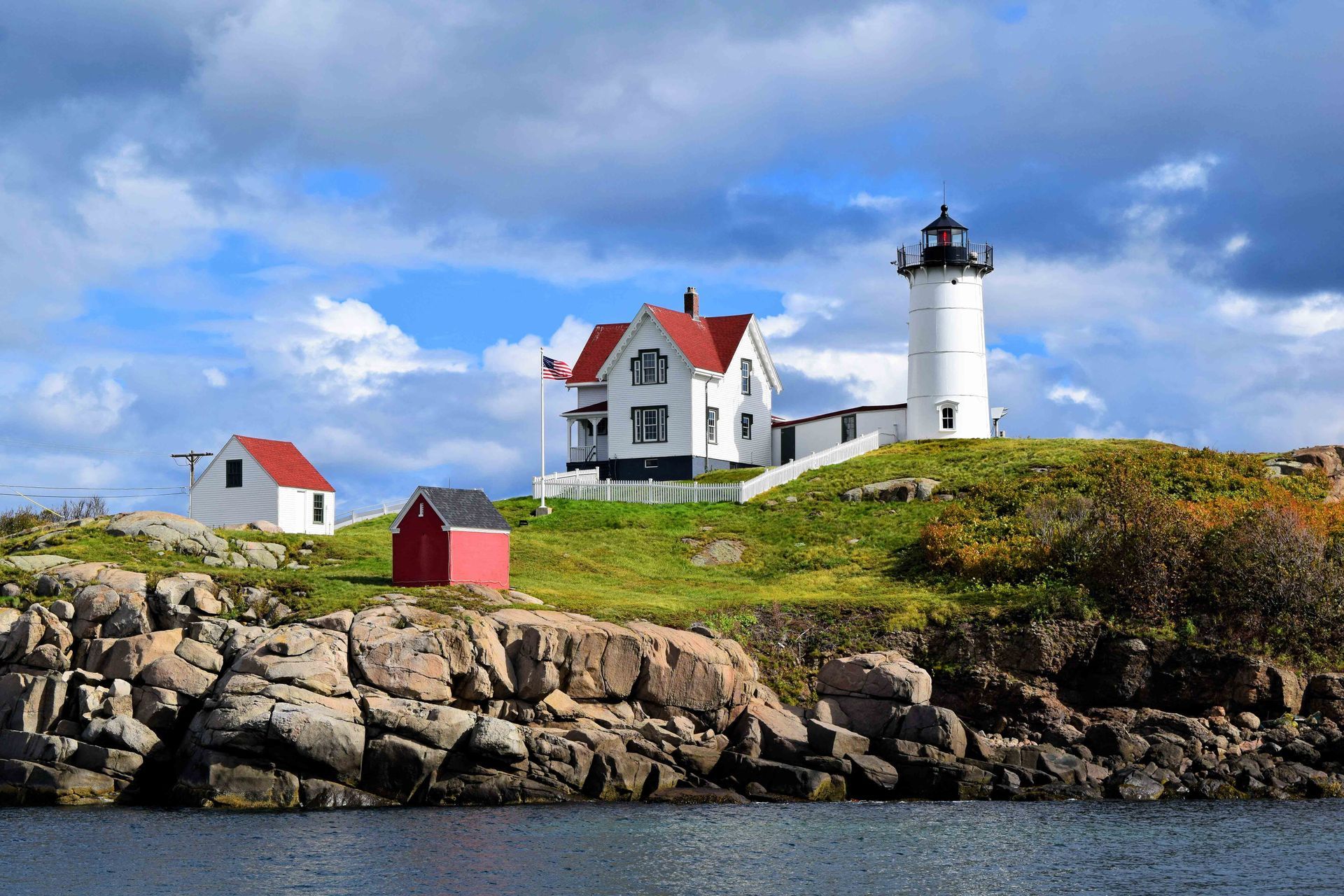 Nubble Lighthouse on a rocky, grassy island under a cloudy blue sky.