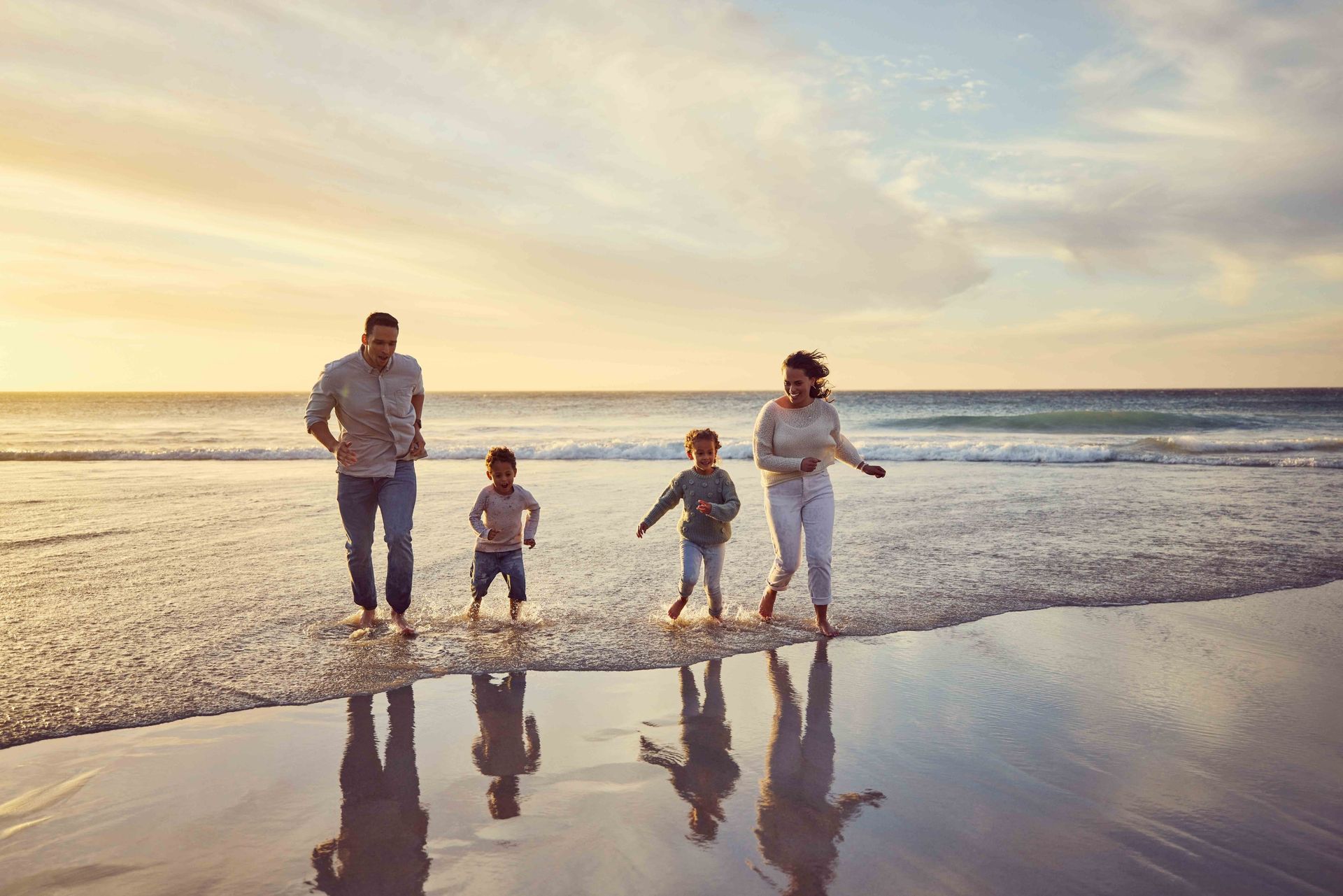 A family of four walking through the shallow surf at the beach during sunset, with soft light reflecting off the water.