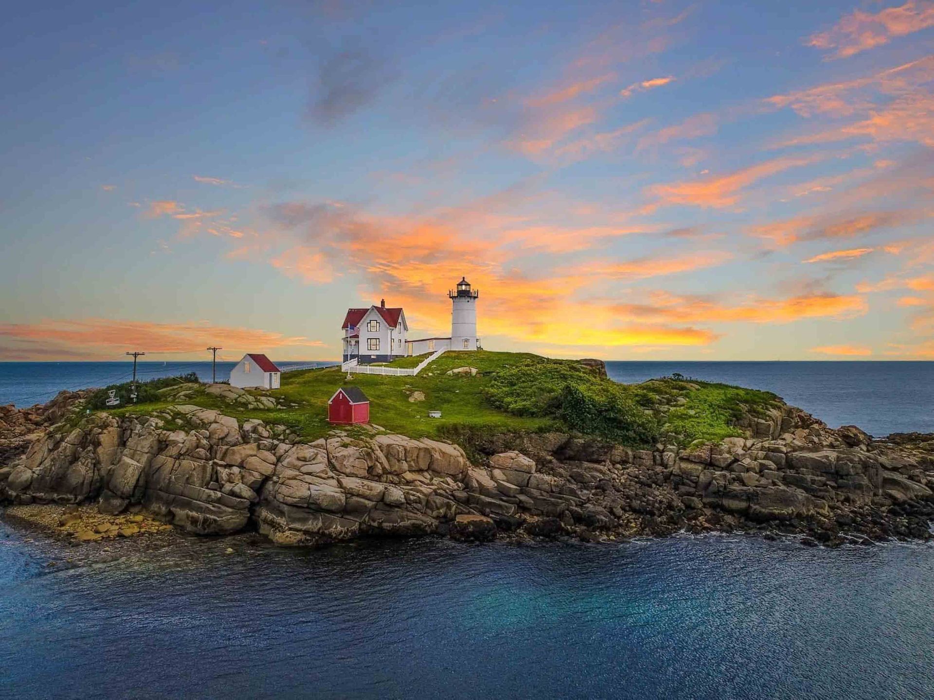 The Nubble Lighthouse sits on a rocky, grassy island surrounded by blue water under a sunset sky with orange clouds.