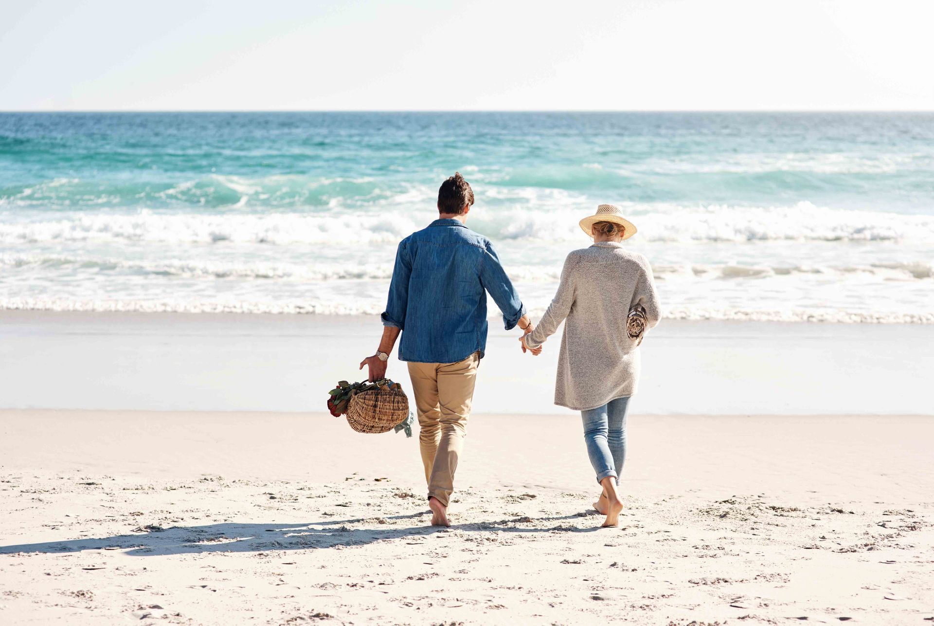 A couple walking hand-in-hand along a sandy beach, carrying a picnic basket toward the ocean.