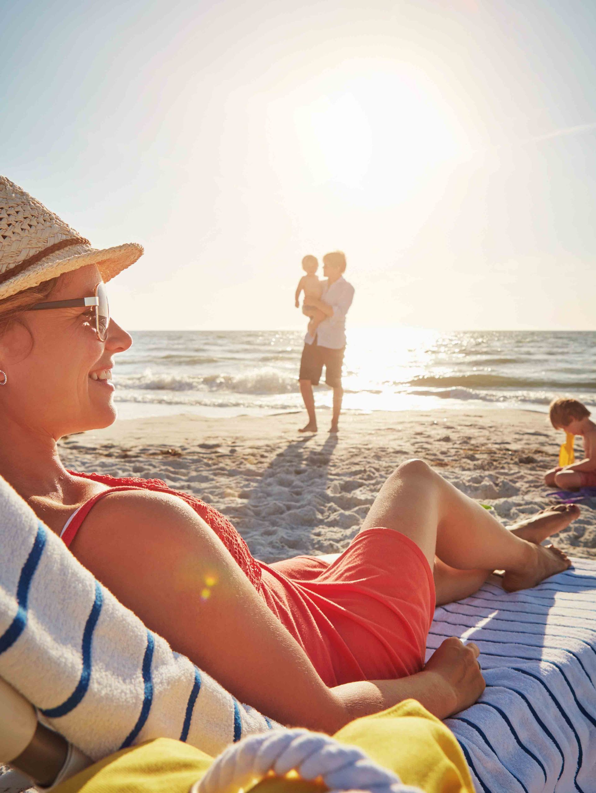 A person in a hat and orange dress relaxes on a beach chair, with a person holding a child and a playing child nearby.