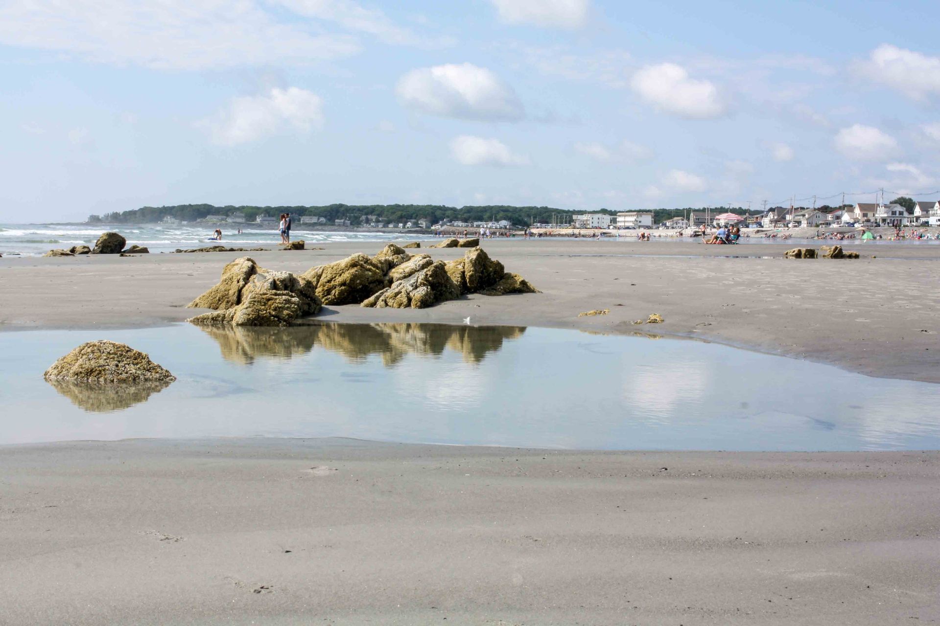 A coastal beach scene with tide pools, rocky outcrops, and a distant shoreline under a partly cloudy sky.