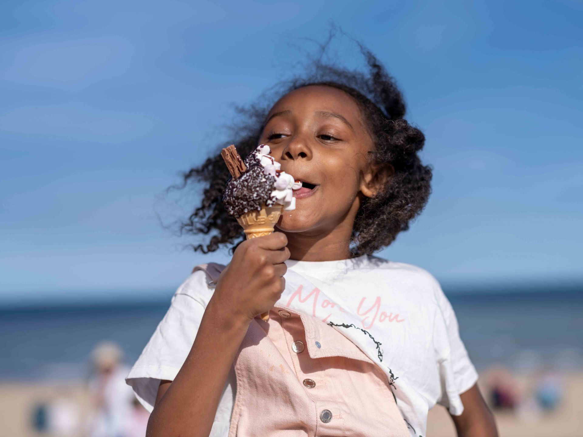 A person eats a chocolate-topped ice cream cone against a backdrop of a blue sky and beach.
