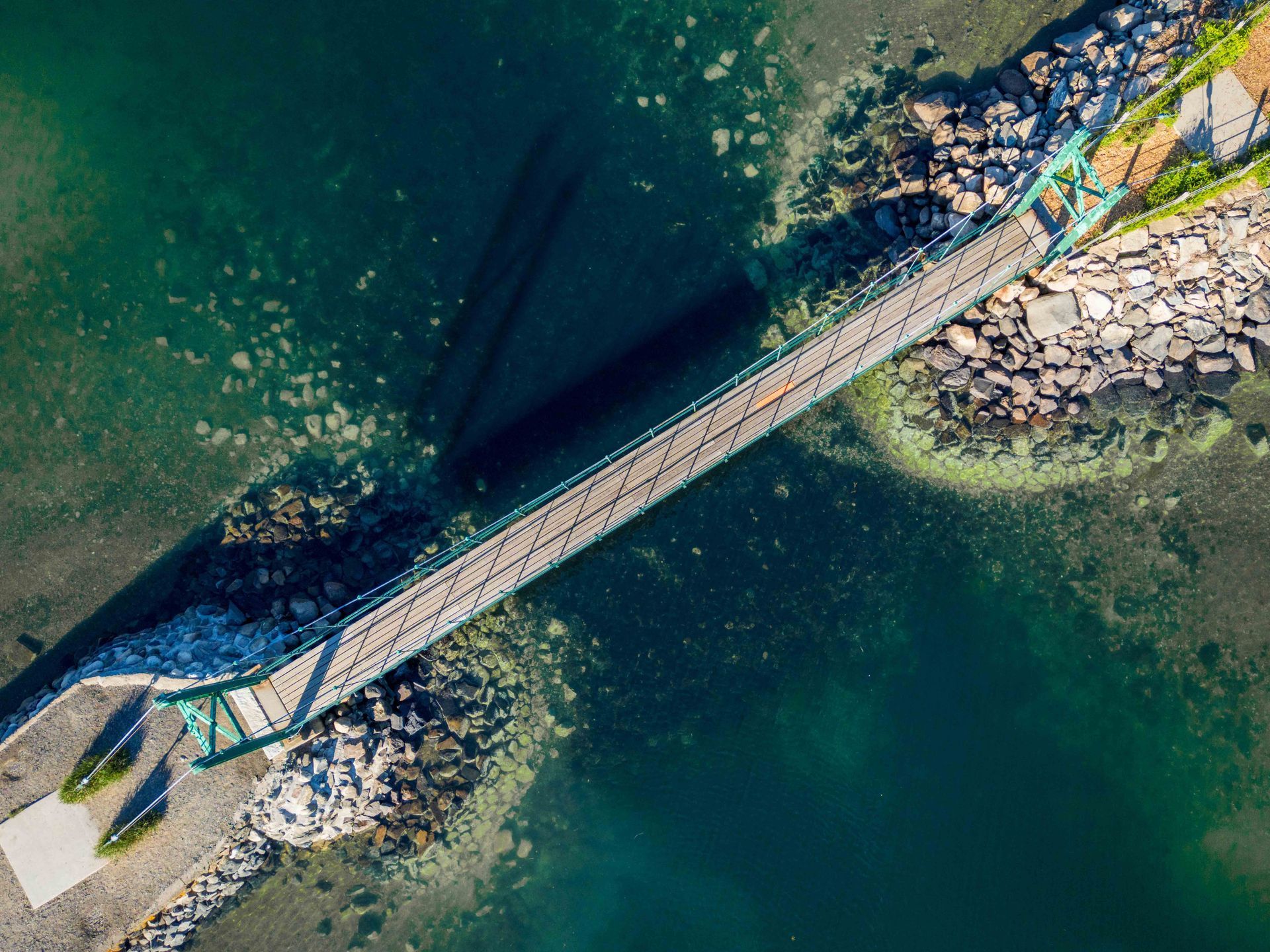 An aerial view of a pedestrian footbridge crossing turquoise water, connecting rocky shorelines on either side.