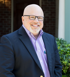 Smiling bald man in glasses wearing a dark blazer and purple shirt outdoors