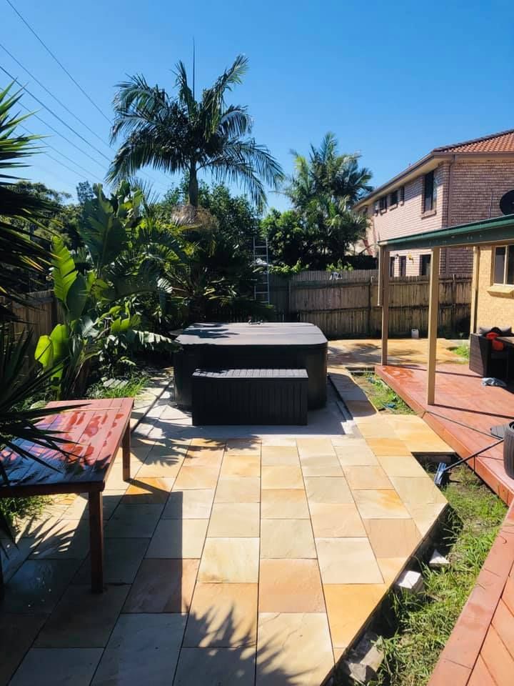 Backyard with hot tub on stone patio, wooden table, lush green plants, and a sunny sky. — East Coast Paving in Seaham, NSW