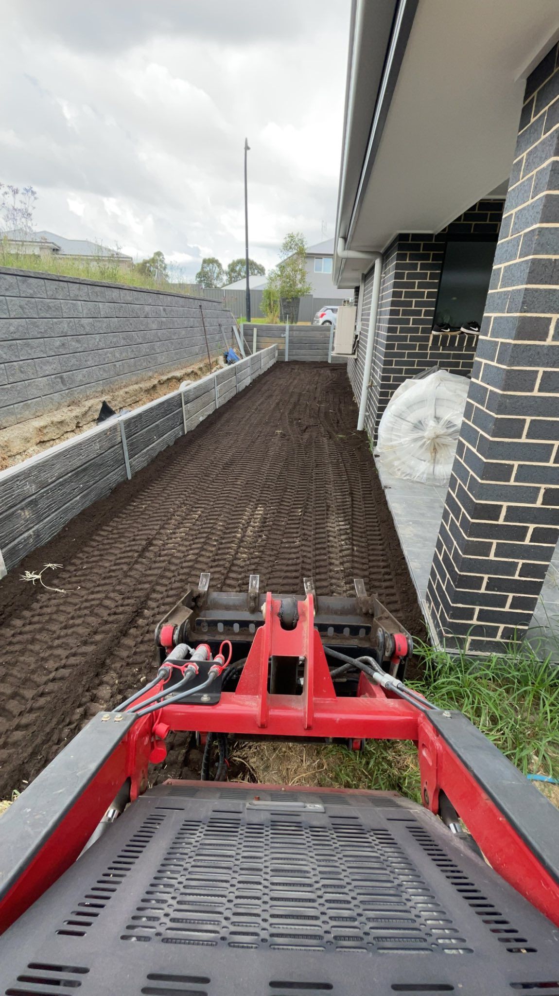 A Red Compact Tractor Tills Freshly Turned Soil Beside a Modern Building — East Coast Paving in Medowie, NSW