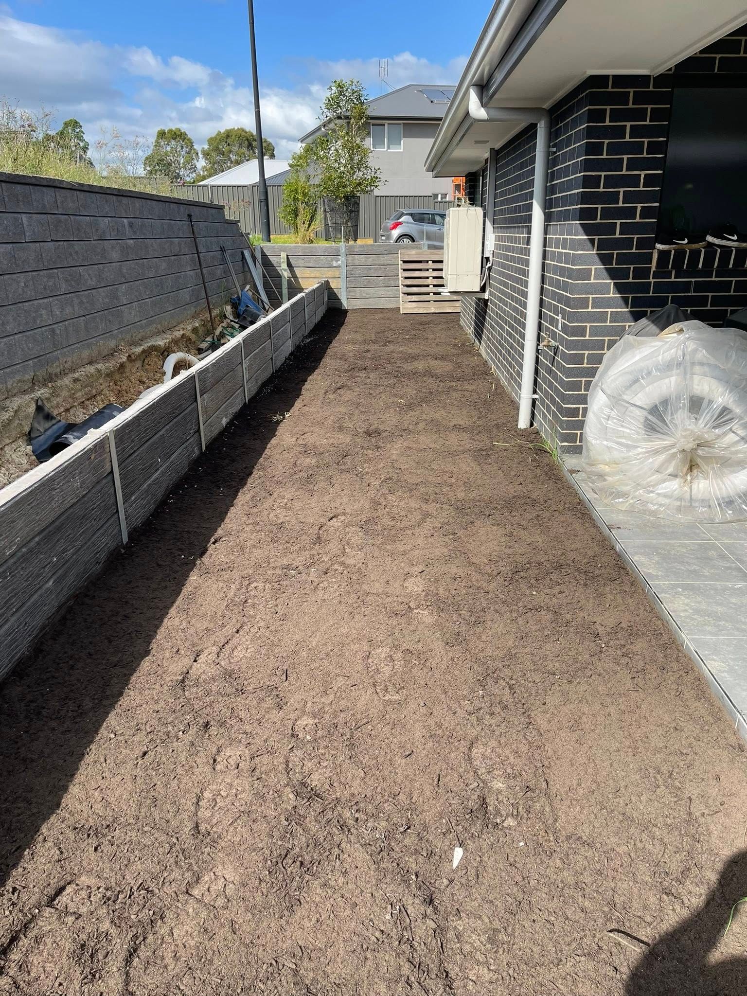 Side yard with fresh soil next to a dark brick wall and a retaining wall on the left. — East Coast Paving in Seaham, NSW