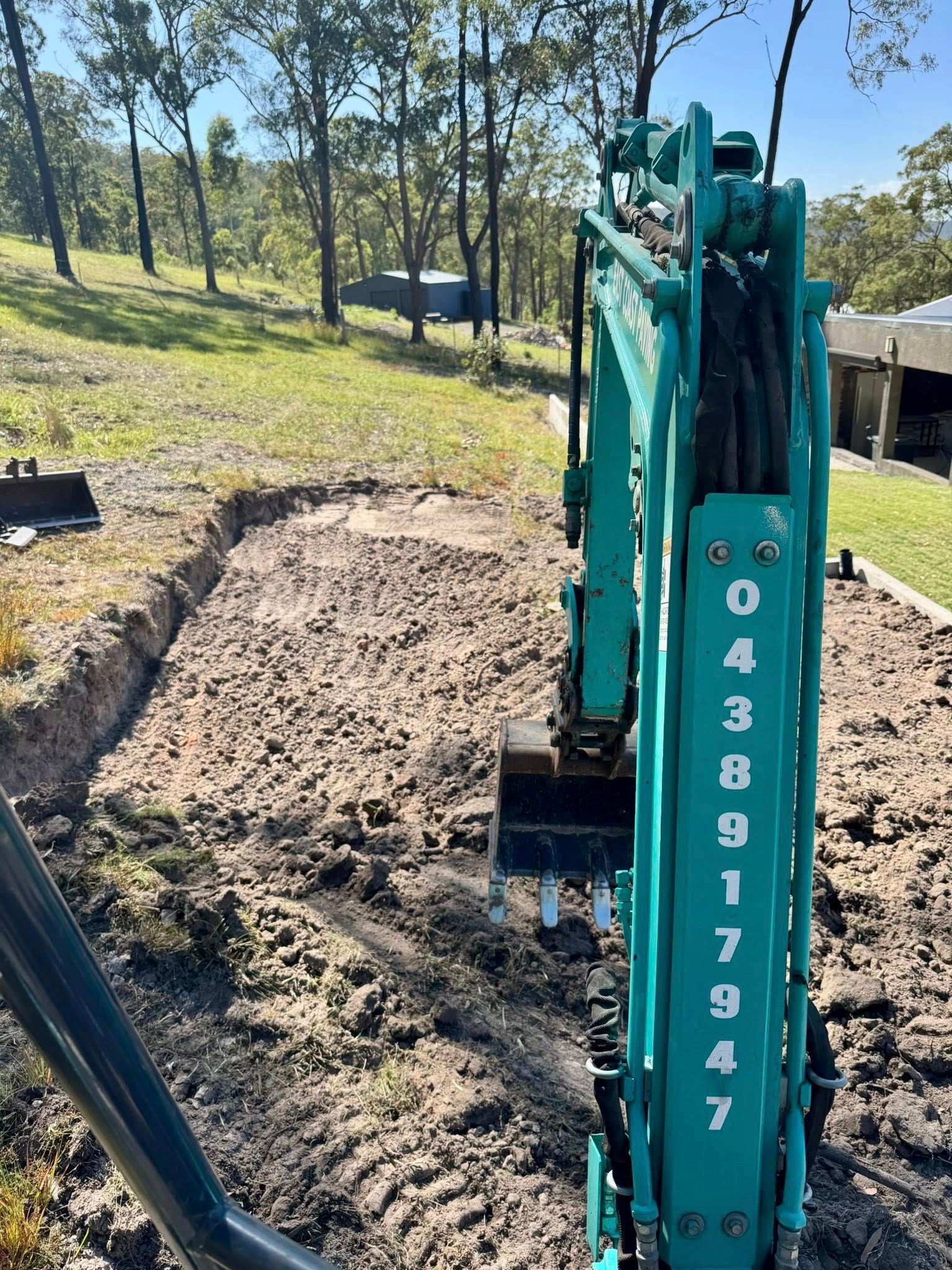 Mini excavator digging a trench in a grassy yard, with a forest in the background — East Coast Paving in Seaham, NSW