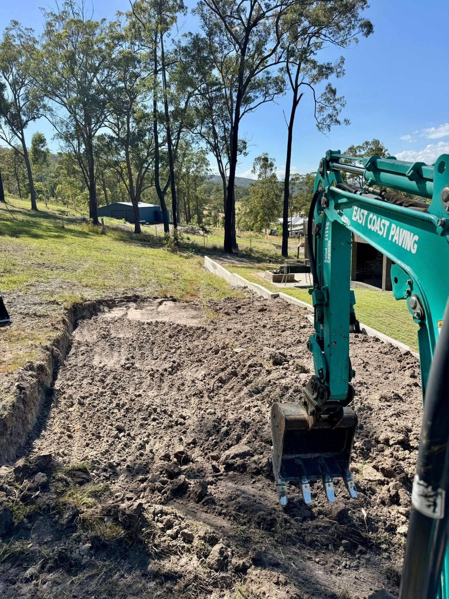 An excavator digging into dirt in a grassy area with trees in the background — East Coast Paving in Seaham, NSW