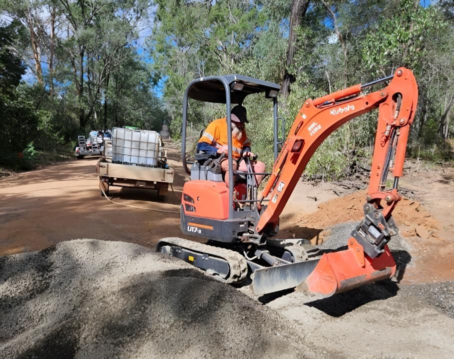 A Man Is Driving A Small Excavator On A Dirt Road — Greenslade's Dozer & Excavator Hire Pty Ltd In Taabinga, QLD