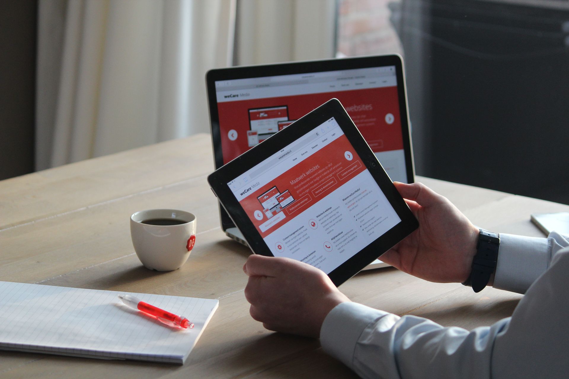 Person holding a tablet at a desk with a laptop, cup, and notebook nearby