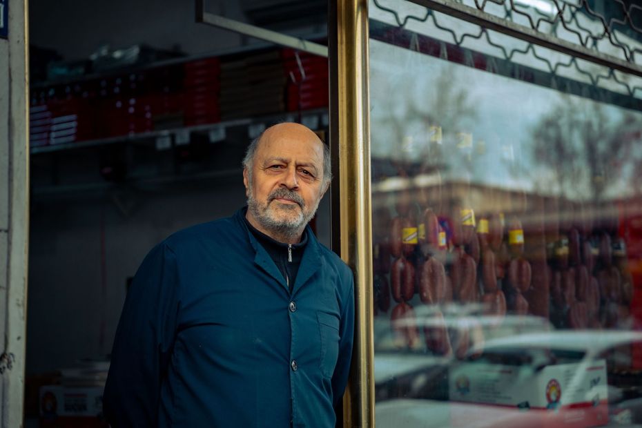 Man standing in a doorway, wearing a teal jacket, with a street reflection in the window beside him.