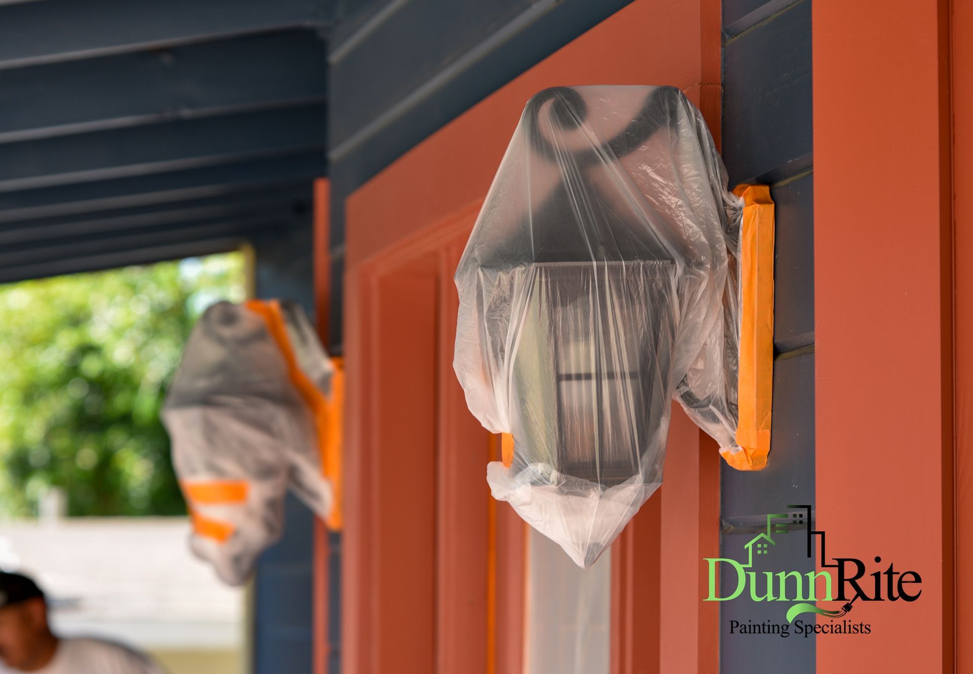 Exterior lights covered in plastic during house painting, orange and blue hues.