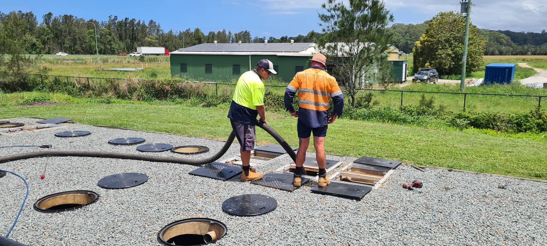 Two Workers, One Holding a Hose, Servicing Septic Tanks in a Gravel Area Near a Green Building  — Raptor Waste Management in Coolangatta, QLD