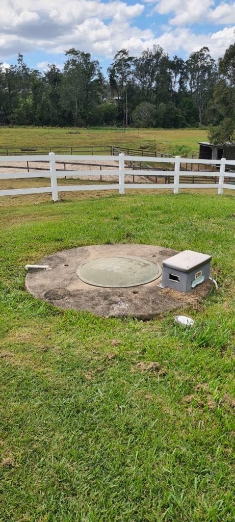 Concrete septic tank lid in a grassy yard, with a small utility box beside it, white fence and trees in background.  — Raptor Waste Management in Coolangatta, QLD