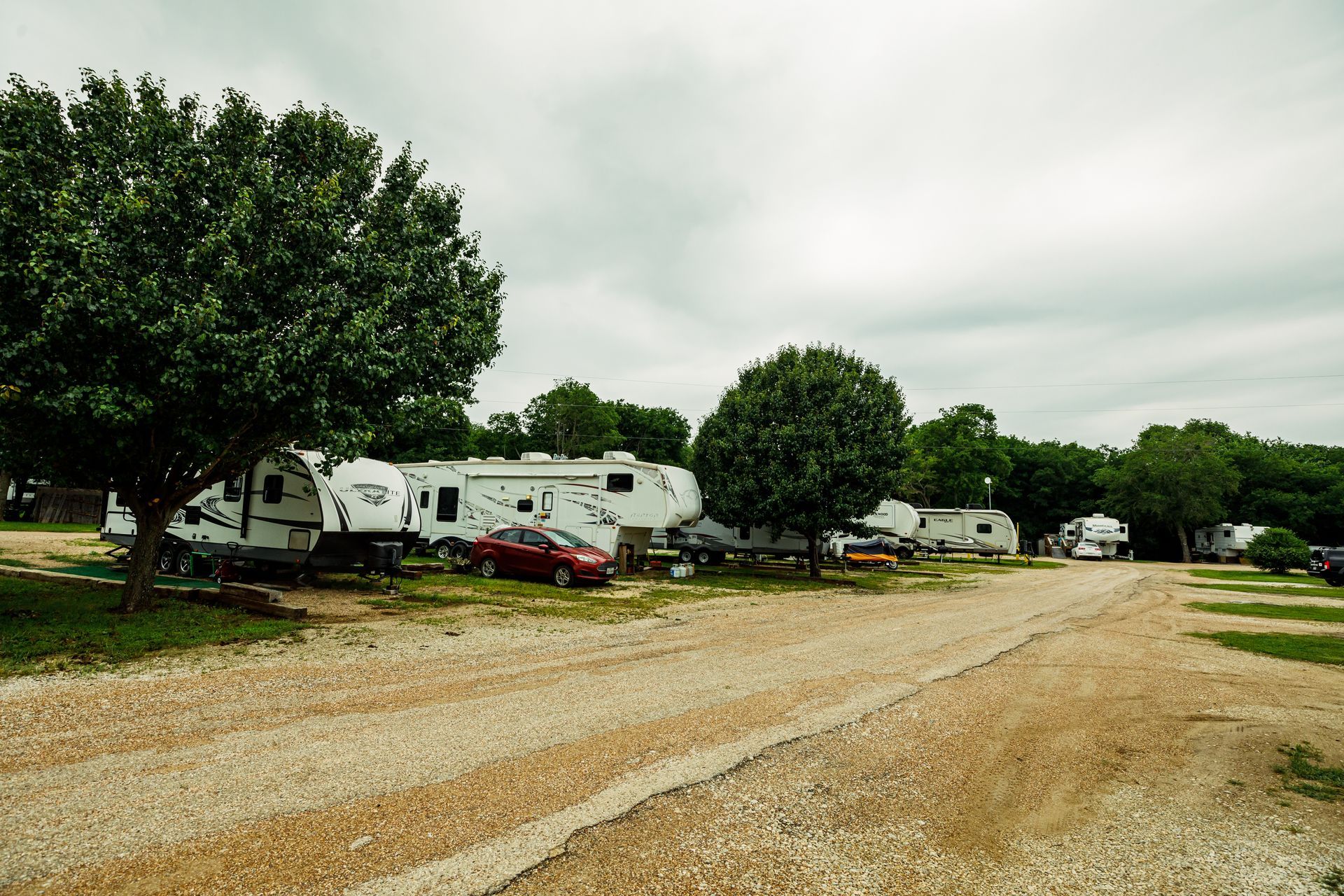 Row of white RVs and cars parked on a gravel lot under cloudy skies, with trees lining the area
