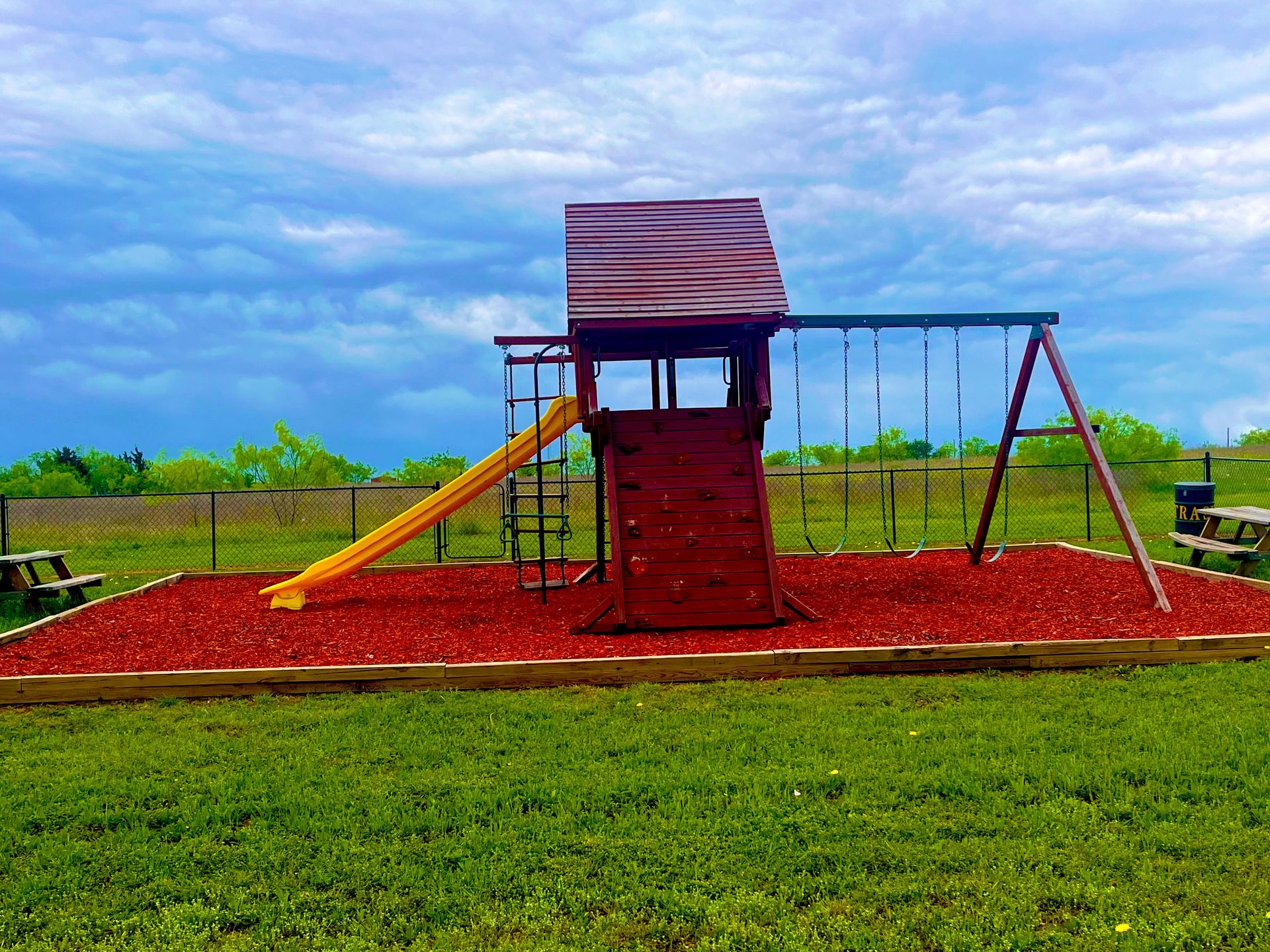Red playground climbing structure with slide and swings on a grassy field under a cloudy sky