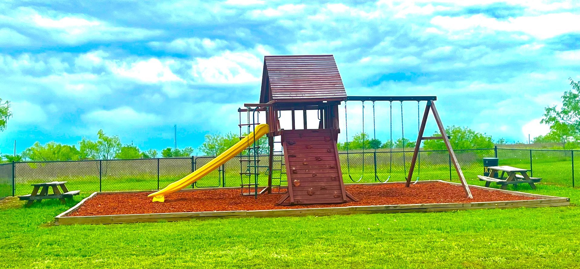 Wooden playground structure with slide, swings, and ladder in a grassy field under a bright blue sky