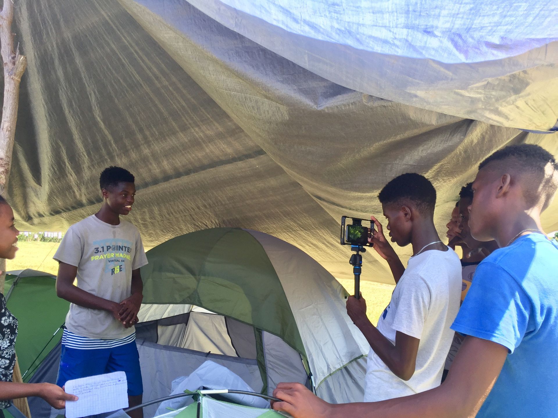 A group of young men are standing under a tarp in front of a tent creating a video.