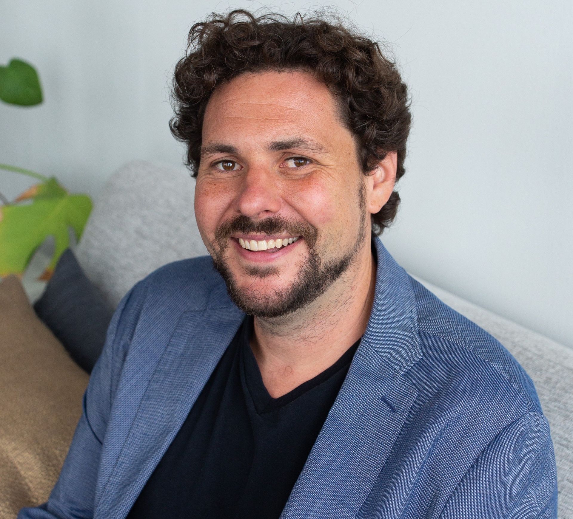 A man with curly hair and a beard is smiling while sitting on a couch. sam mestman