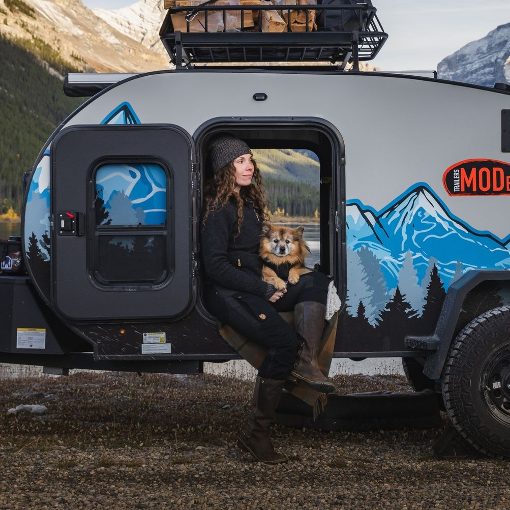 Woman with dog sitting in the doorway of a gray camper van in the mountains