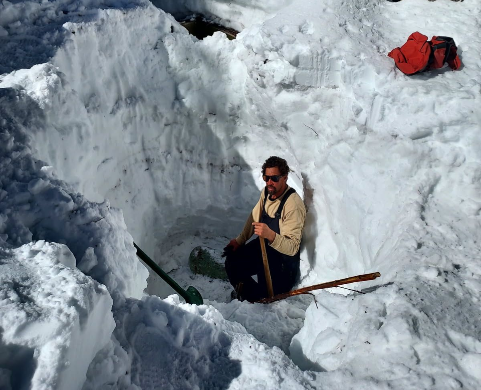 Man in overalls and sunglasses kneels in a snow pit, holding a shovel. Red jacket rests nearby.