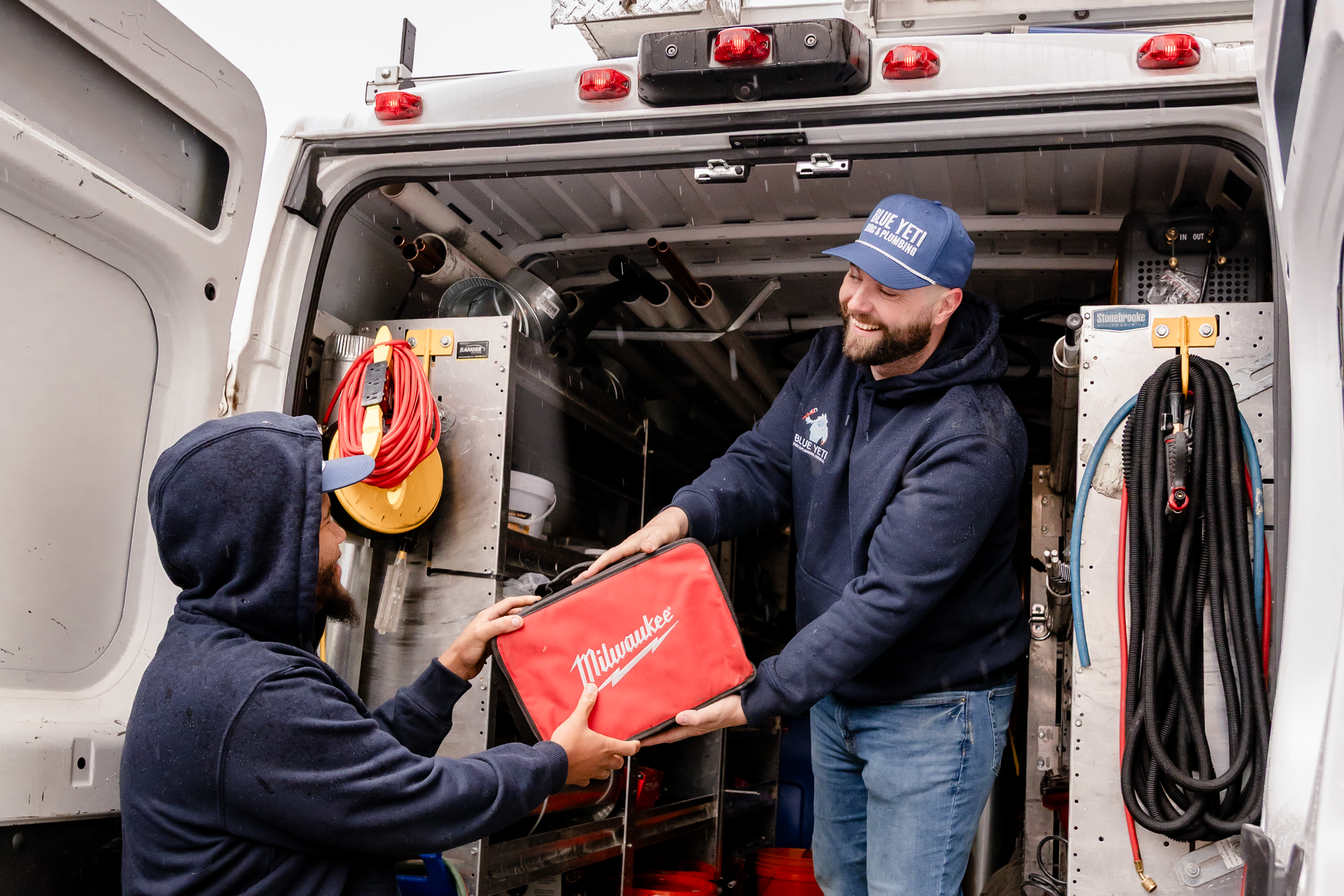 Two Blue Yeti HVAC Plumbing technicians exchanging a red tool bag inside a utility van