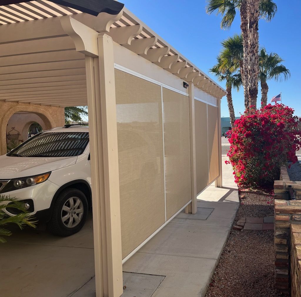 Beige sun shades on a carport with a white car parked inside. Palm trees and red bush in the background.