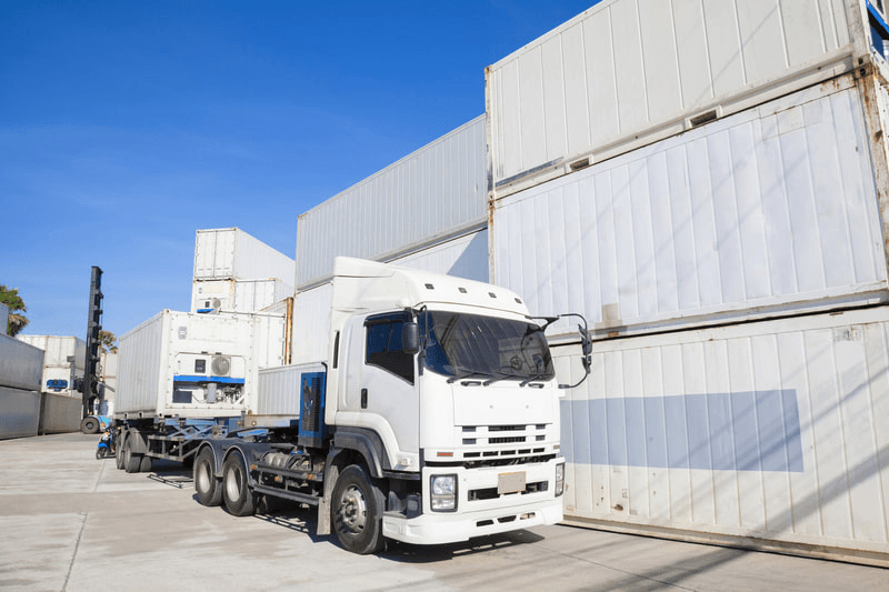White semi-truck hauling shipping container, parked at a container yard on a sunny day.