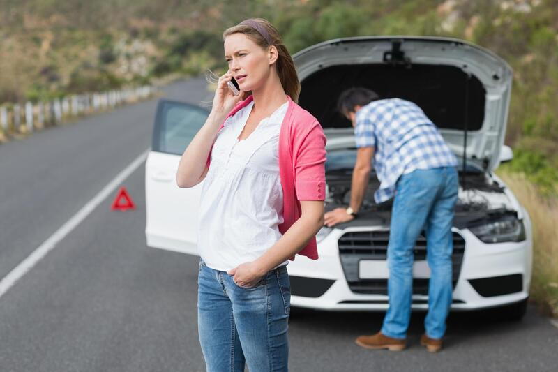 Woman on phone beside a broken-down white car on a road; man looks under the hood; a red warning triangle is on the road.