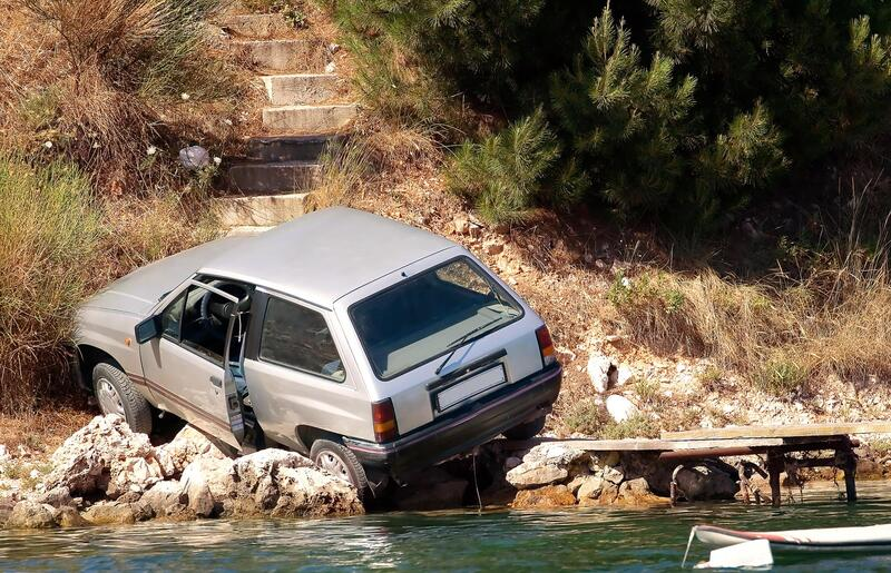 Silver car crashed near water, with door open, next to small dock and stone wall, outdoors.