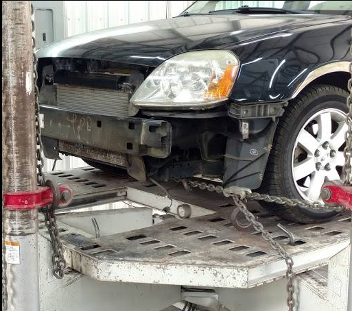 A woman is standing in front of a damaged car and looking at her phone.