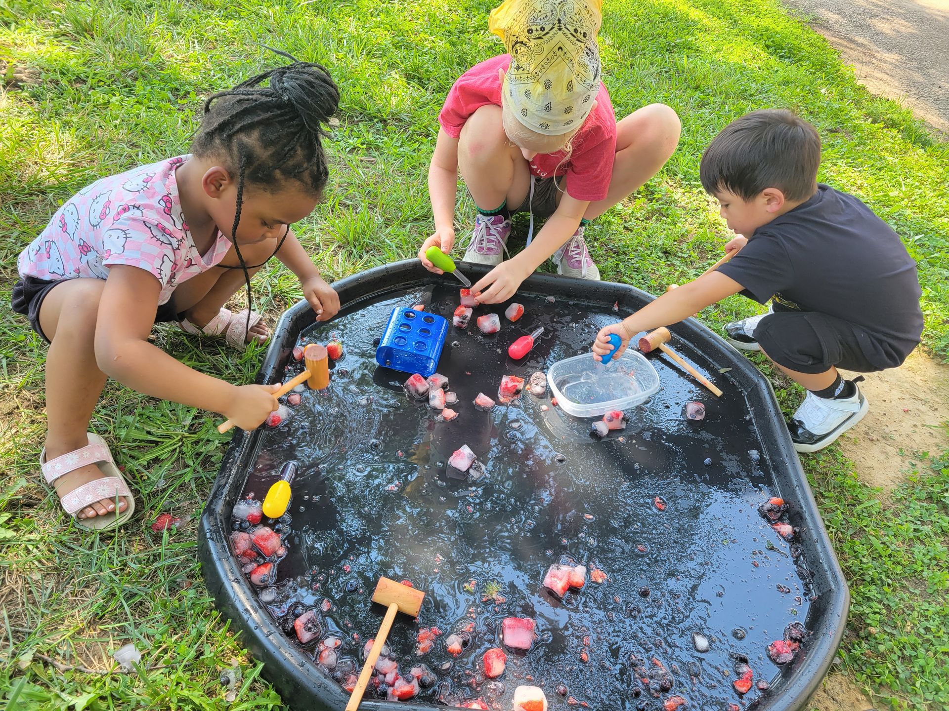 The Seedlings Outdoor Learning program provides access to nature-based play with the Association for Nature-Based Education. Here, three children crouch to play with water, crab mallets, and natural loose parts in a sensory table outdoors.
