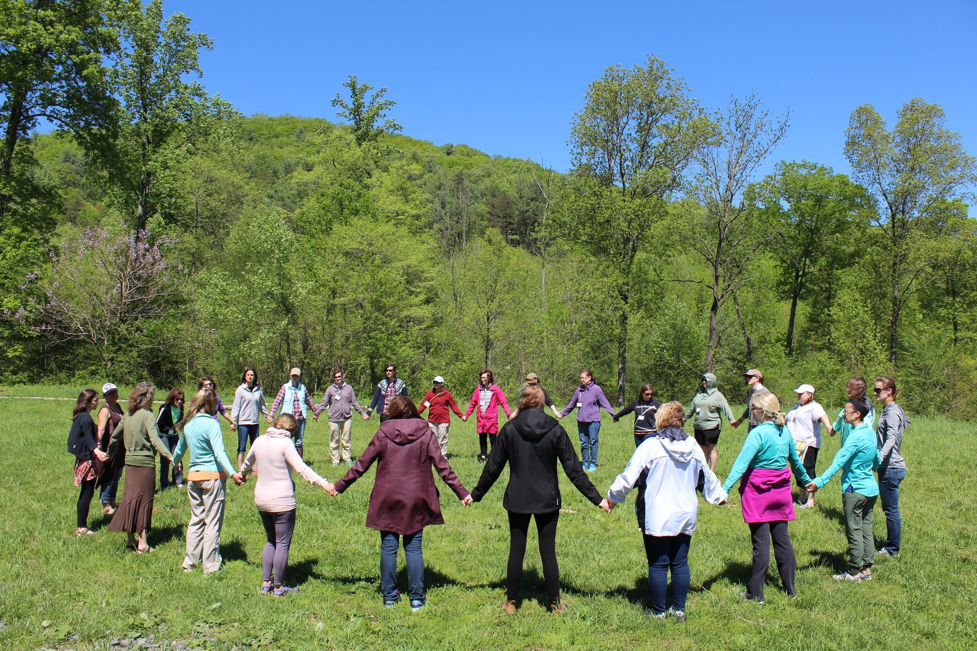 A group of about 30 teachers hold hands in a circle outdoors as they gather for professional development with the Association for Nature-Based Education.