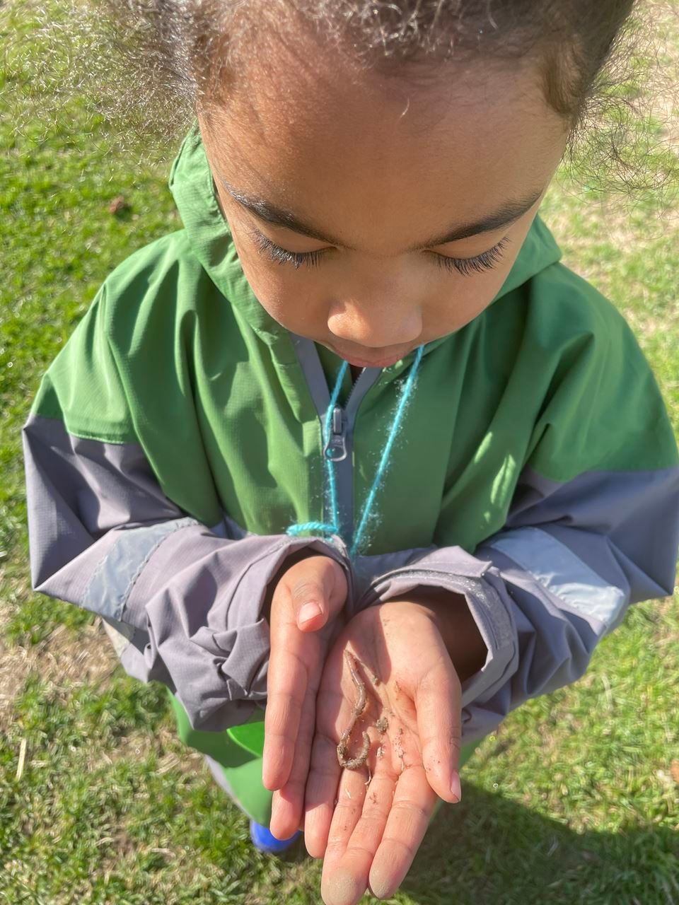 Kindergarten child wears a rain suit outside and holds hands open to share a worm as part of the Schoolyard Outdoor Learning Experiences (SOLE) Program hosted by the Association for Nature-Based Education.