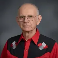Elderly person in glasses wearing a red and black patterned shirt against a gray backdrop