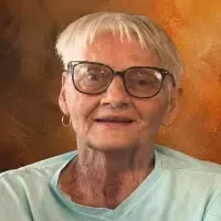 Portrait of an older person with glasses and short white hair, wearing a light teal shirt against an orange background